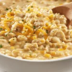 Angled hero shot of creamy white chicken chili in a white bowl with shredded chicken, white beans, green chiles, melted Monterey Jack, cilantro, and tortilla strips, with a spoon and linen napkin