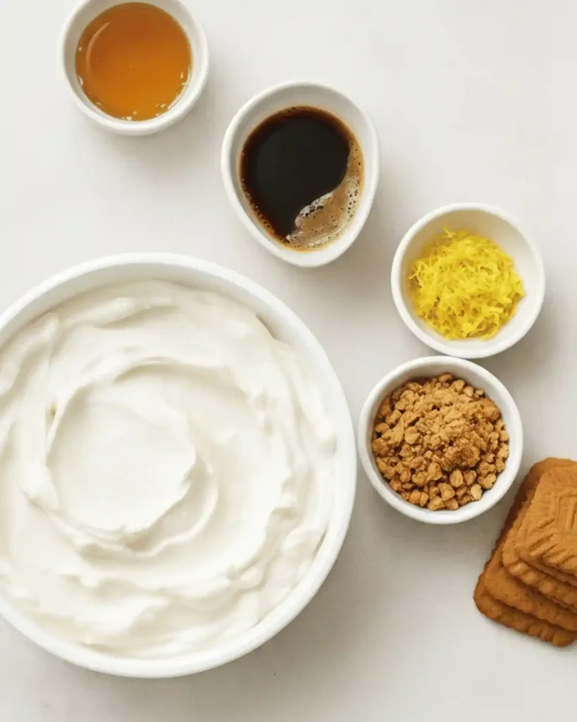 Plain Greek yogurt and Biscoff cookies arranged on a light surface with small bowls of optional honey, vanilla, lemon zest, and cookie crumbs.